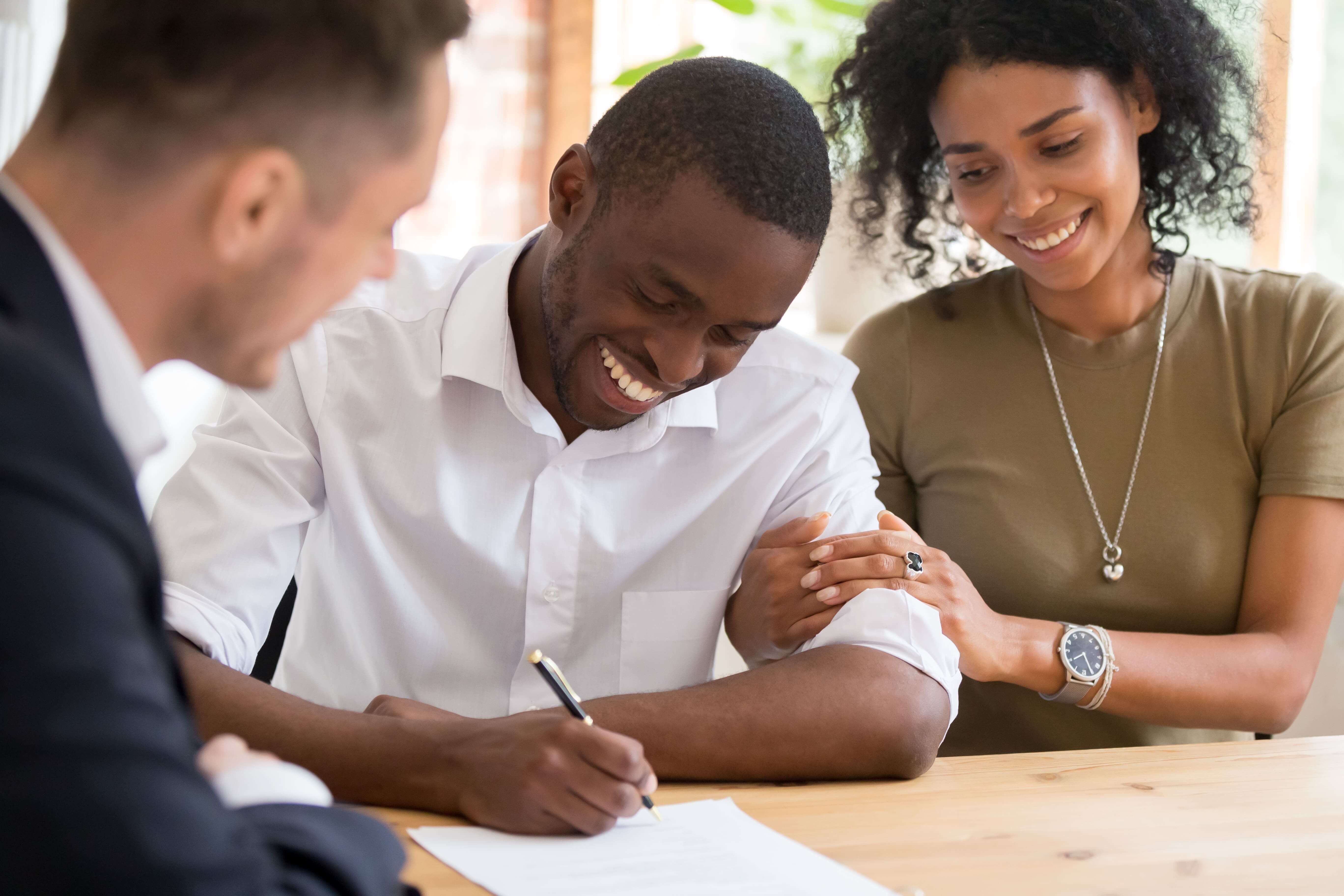A smiling man holds a pen in his hand and signs a document while a woman clutches his arm and a man looks on. 