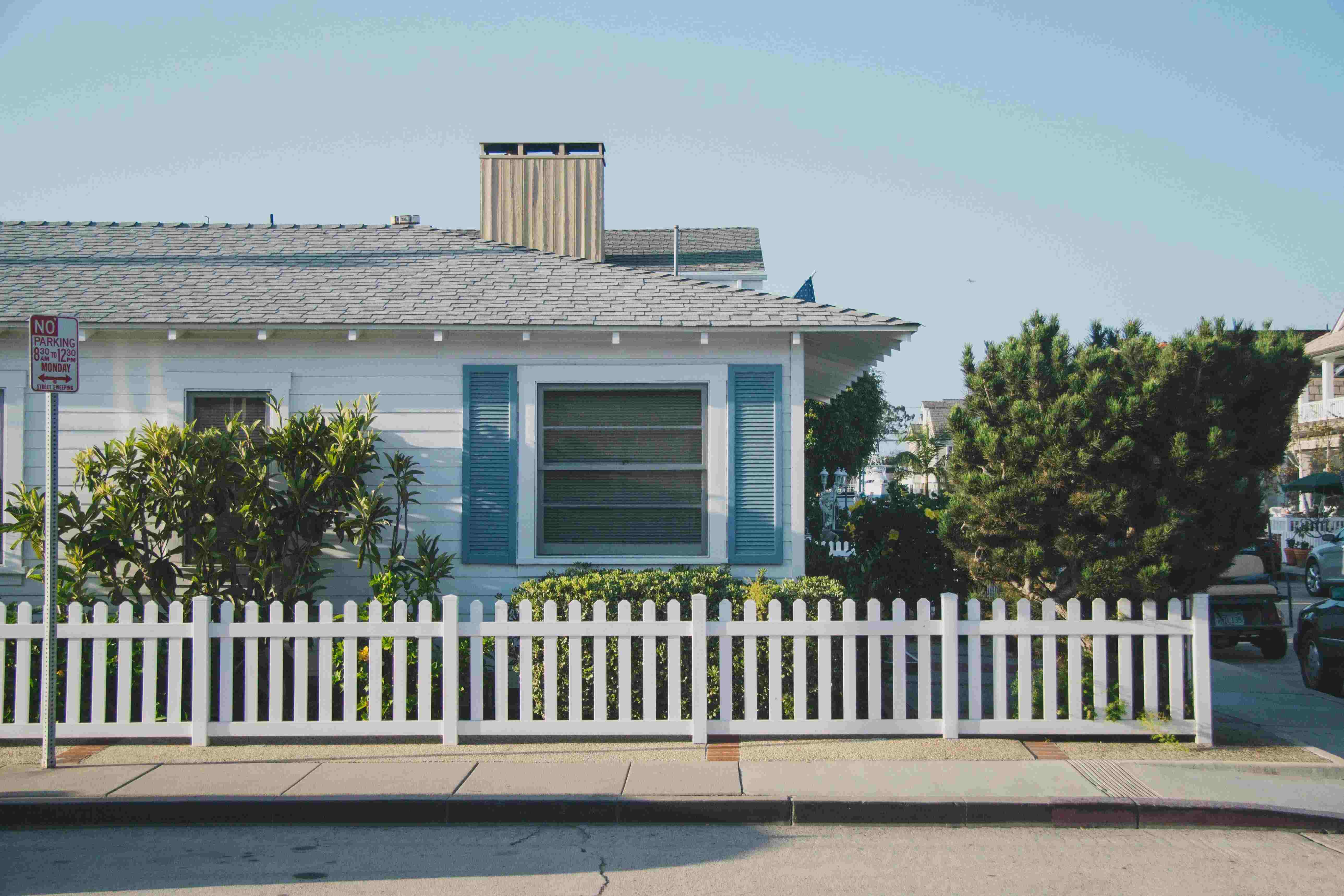 White house with blue shutters and a white picket fence with lush green bushes and two medium sized trees. 