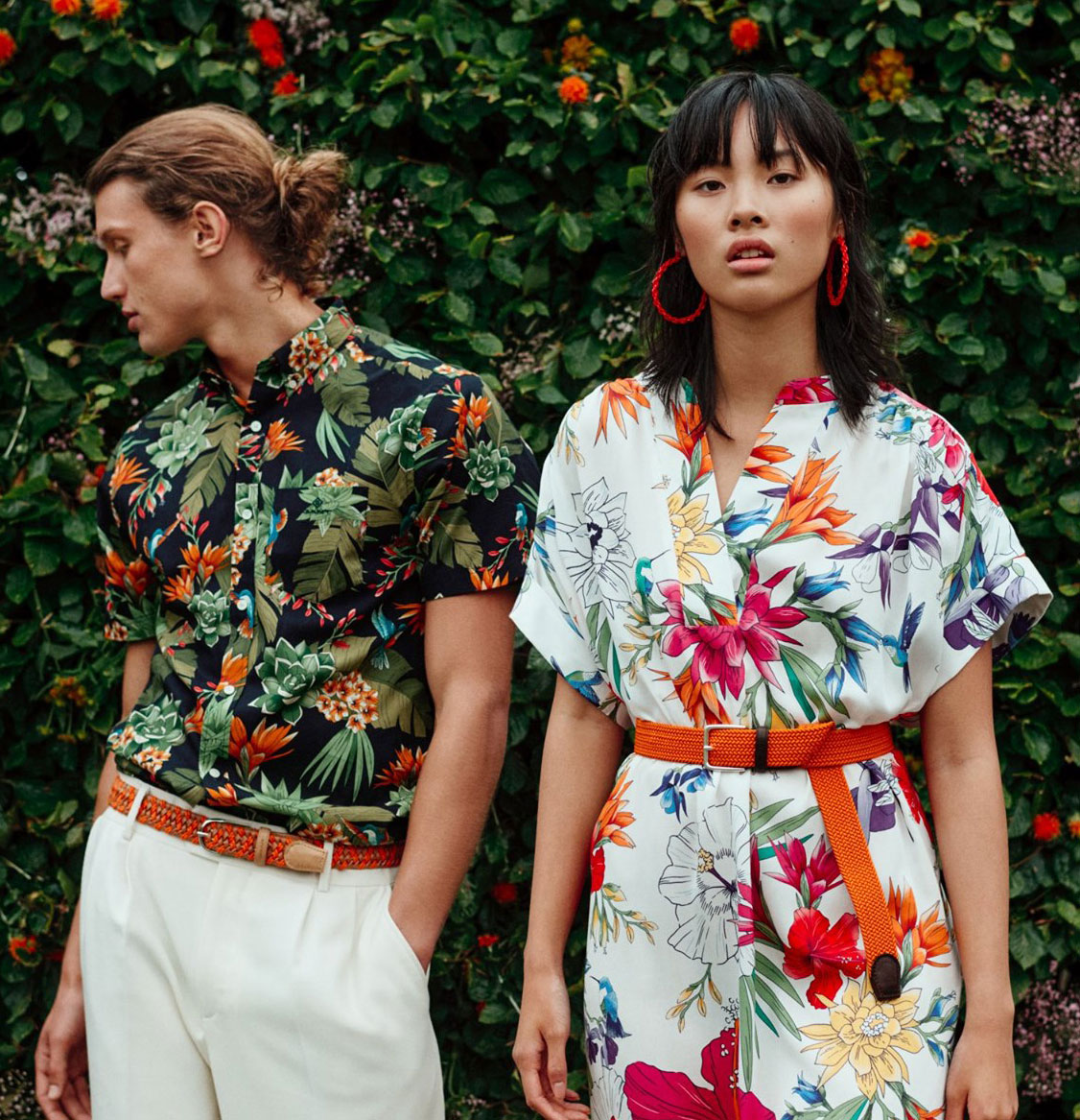 Two models stand wearing bold, tropical print outfits with red accessories.