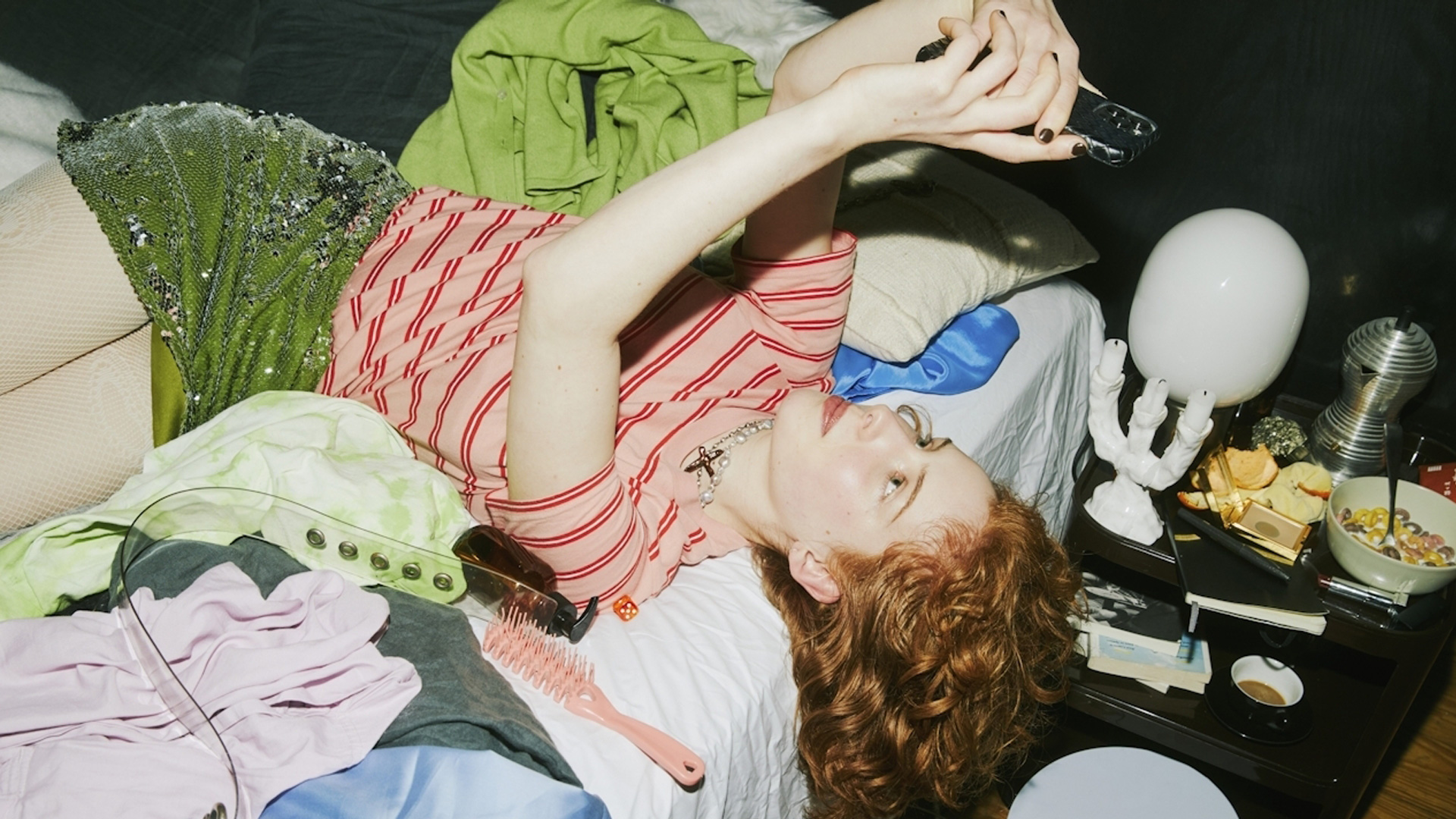 Woman with red hair in a striped top and green sequin skirt, lying on a messy bed looking at her phone.