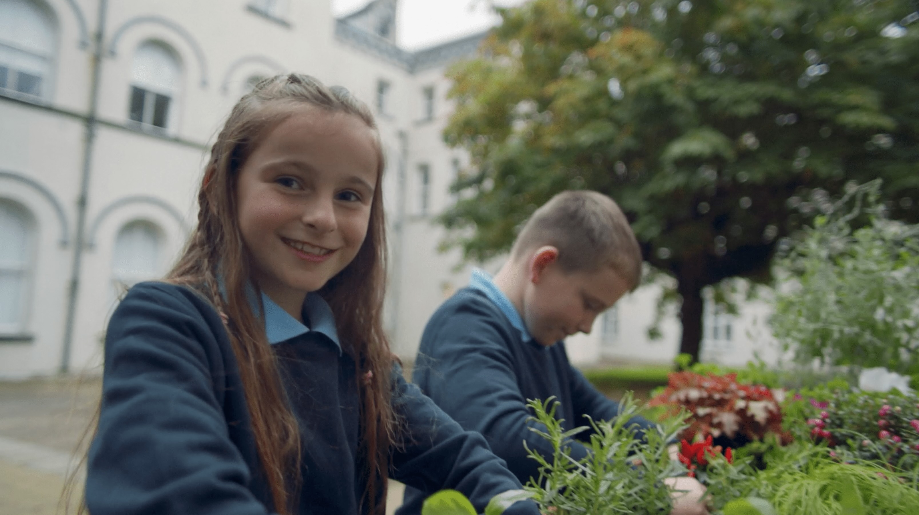 A young girl smiling while tending to a garden, with another child working in the background. The setting appears to be an outdoor school courtyard with white building arches in the background.
