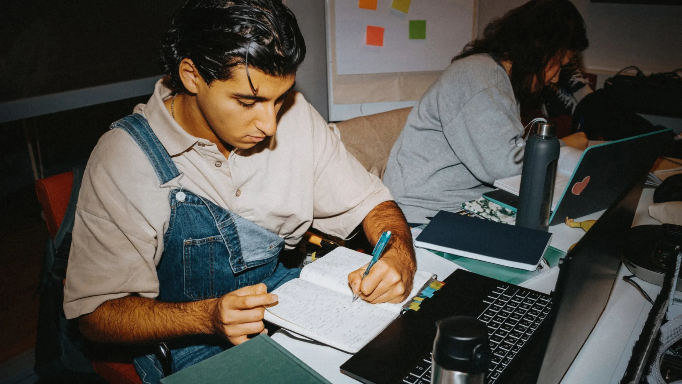 A warm, indoor photo showing two young adults studying together at a desk. The person in the foreground wears denim overalls over a beige shirt and is focused on writing notes in a physical notebook with a pen. The second person is seated behind, looking down at a laptop.